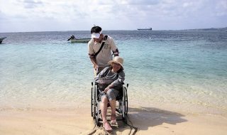 ヘルパーと旅する笑顔の車いすの女性。砂浜には車輪の跡。バックには美しい海が。